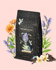 A black bag of Vanilla Lavender Black Iced Tea is surrounded by lavender, orange flowers, and vanilla pods, set against a light pink background. The whimsical packaging features stars and birds, capturing the calming essence of lavender and the rich depth of organic Assam black tea.