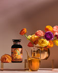 A decorative display featuring "Venus - Tea for Beauty Sleep" in a bottle with vibrant artwork, complemented by a colorful floral arrangement in a gold vase and a glass carafe filled with golden liquid. The setup is placed on a neutral-toned backdrop, accentuating the vivid botanicals of the flowers and the detailed art on the bottle.