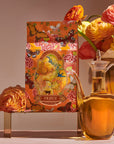 A floral-themed product package labeled "Venus - Tea for Beauty Sleep" with bright, colorful illustrations is displayed alongside a clear glass jug of golden liquid, possibly oil. In the background, a copper vase is filled with vibrant, multicolored ranunculus flowers and botanicals, creating a warm, inviting scene.