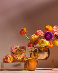 A glass jug of Venus - Tea for Beauty Sleep sits next to an orange floral perfume box. A shiny gold vase filled with vibrant, multi-colored ranunculus flowers is placed on a raised stand behind them. The background is gradient beige, creating a warm, botanicals-inspired atmosphere.