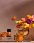 A stylish still life composition features a metallic vase filled with vibrant, colorful ranunculus flowers, a sleek glass decanter with amber liquid, and a small cylindrical container of Venus - Tea for Beauty Sleep with elaborate artwork, all set against a warm, neutral background.