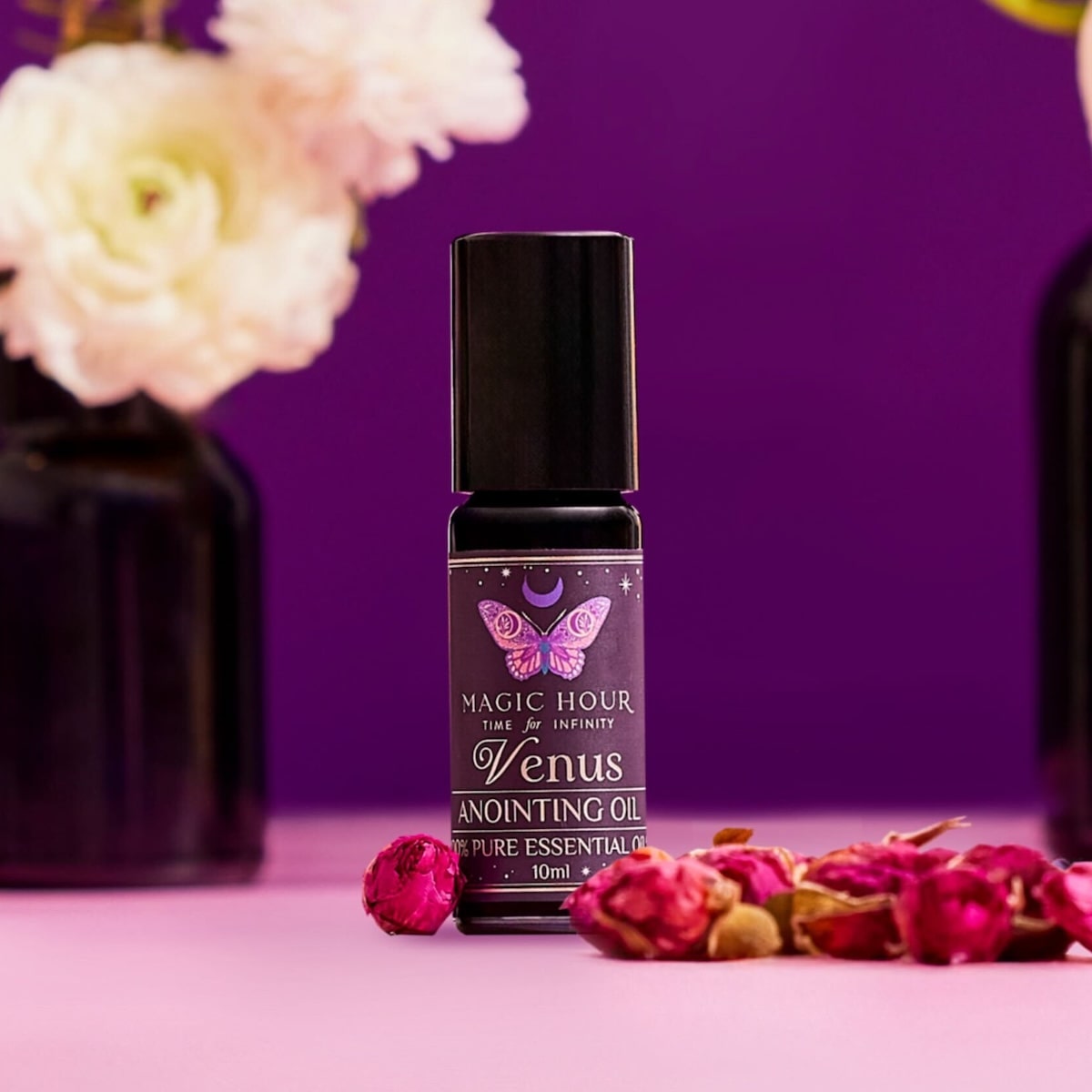 A bottle of Planetary Perfume Oil, made with pure essential oils, rests on a pink surface with dried rosebuds, blurred white flowers, and a purple background.