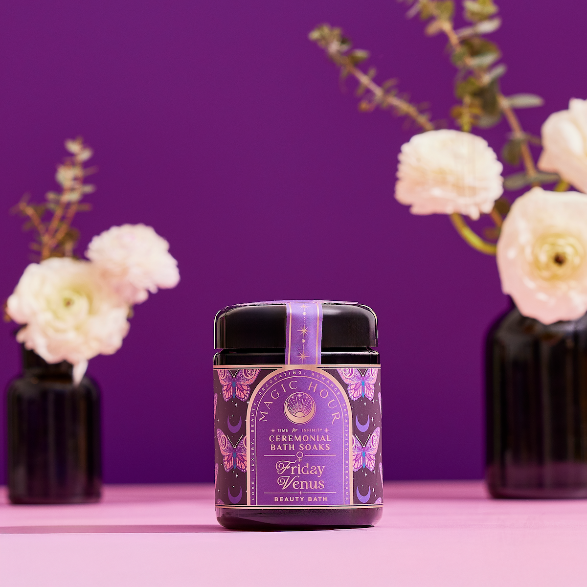 A jar of Ceremonial Bath Salt Soaks - Violet Glass Jar with a purple and gold label sits on a pink surface, creating a celestial vibe, while white flowers in black vases stand against a purple background.