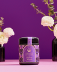A jar of Ceremonial Bath Salt Soaks - Violet Glass Jar with a purple and gold label sits on a pink surface, creating a celestial vibe, while white flowers in black vases stand against a purple background.