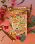 The Magic Hour Starter Kit, prominently featuring the brand's "Venus" tea, is set against a gradient backdrop of pink and orange hues. The bag is encircled by sprigs of red flowers, lavender, and green foliage. Accompanying the kit are two golden measuring spoons engraved with "Magic Hour," ideal for elevating your next tea ceremony.