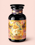 A dark glass bottle adorned with a decorative label featuring flowers, butterflies, and an art nouveau-inspired illustration of a woman. The label reads “Magic Hour Venus - Tea for Beauty Sleep,” hinting at the caffeine-free herbal blend inside, crafted from beautifying botanicals. The bottle is set against a light pink background.
