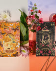 A visually captivating display features two tarot card decks surrounded by flowers and leaves. The left deck, part of Magic Hour's "The Lovers Bundle with Venus + The Lovers Tea," depicts 'Venus' with an orange floral design, while the right deck shows 'The Lovers' with a romantic night sky and floral theme on a gradient background.