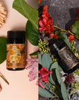 A split image showcases two bottles from Magic Hour's The Lovers Bundle with Venus + The Lovers Tea, nestled among flowers. On the left half, a black bottle with an orange label featuring a woman and floral artwork is surrounded by pink blooms and green leaves. The right half displays a similar bottle adorned with a dark label, set amidst red flowers and green foliage, exuding the allure of sensual flavors.