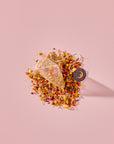 A pyramid-shaped tea bag of Tea for Rest: Venus: Mango Blossom & Nervines Herbal Tea rests on a pile of loose-leaf herbal blend containing dried flower petals, buds, and organic botanicals, with a circular tag attached.