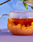 A clear glass mug filled with hot tea from The Best of Fall Box sits on a light purple surface. Steam rises from the Pecan Pie Chai, and a swirling pattern of cream is being mixed into it. A branch with green leaves is partially visible in the background against a blue sky.
