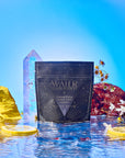 A packet of Water sits on reflective water, surrounded by flowers, lemon slices, and a large crystal, with a bright blue sky in the background.