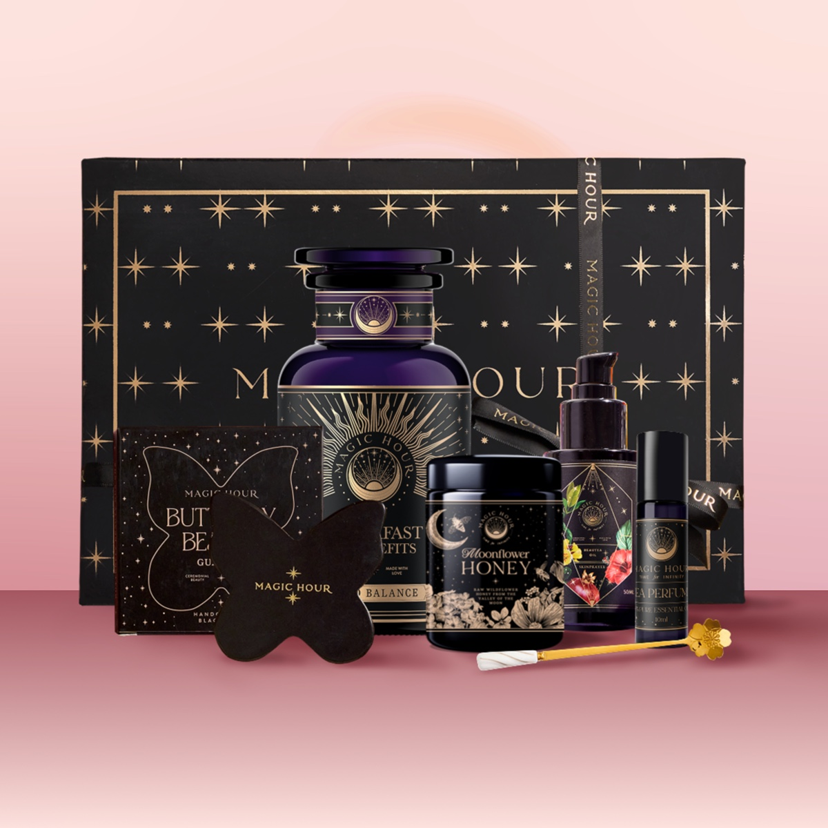 The Wake Up to Beauty Gift Box features Magic Hour tea, honey, a candle, spray bottle, heart-shaped tool, and more in elegant black and gold packaging—perfect for your morning ritual and presented on a pink gradient background.