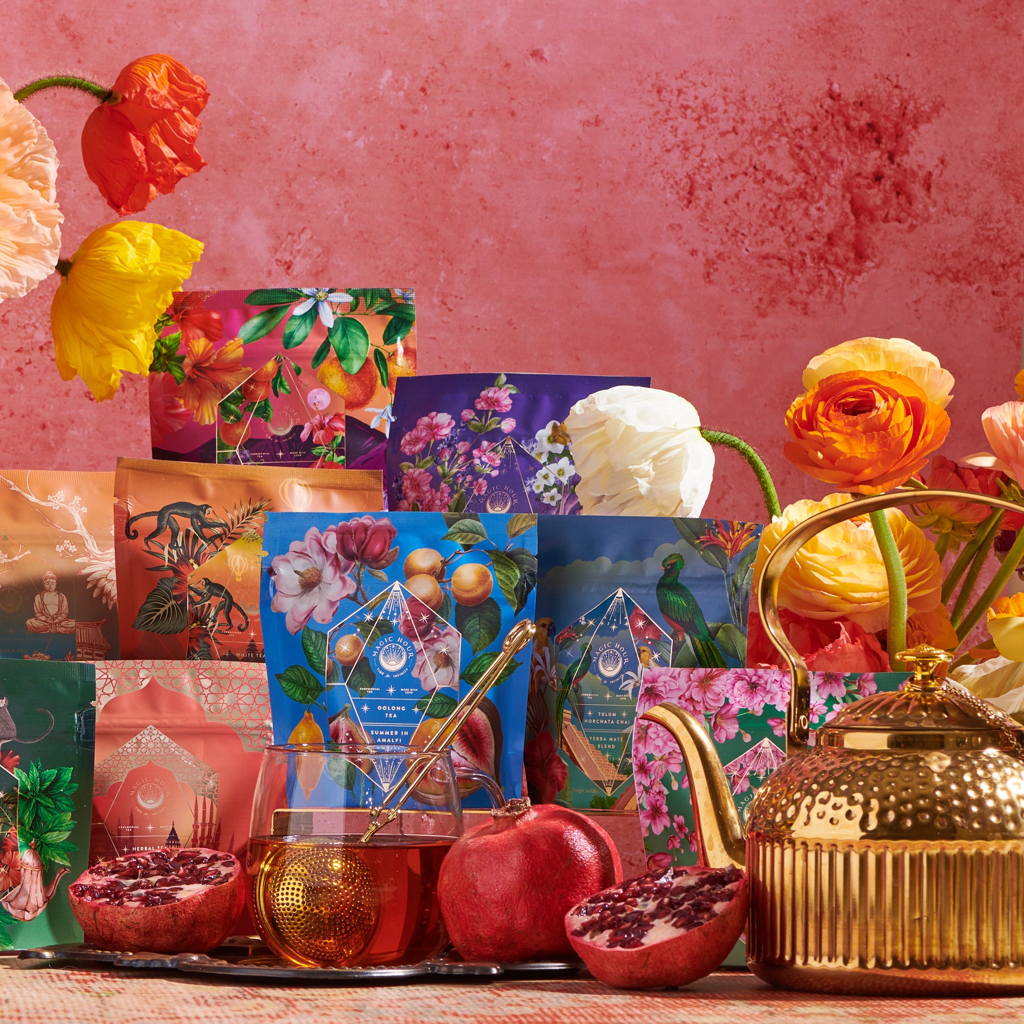A colorful display showcasing the vibrant Wanderlust Sampler Tea Set, featuring floral and botanical designs, arranged against a pink wall. The scene includes blooming flowers, fresh pomegranates, a glass honey pot with a honey dipper, and a golden teapot.