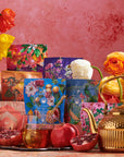 A colorful display showcasing the vibrant Wanderlust Sampler Tea Set, featuring floral and botanical designs, arranged against a pink wall. The scene includes blooming flowers, fresh pomegranates, a glass honey pot with a honey dipper, and a golden teapot.