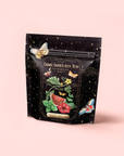 A black resealable pouch labeled "Watermelon Mint Herbal Iced Tea" displays watermelon, mint leaves, red flowers, butterflies, and stars on a light pink background—featuring organic herbal tea with watermelon juice powder.