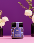 A dark violet glass jar of Ceremonial Bath Salt Soaks rests on a pink surface, exuding celestial healing energy, with white flowers in black vases against a vibrant purple backdrop.