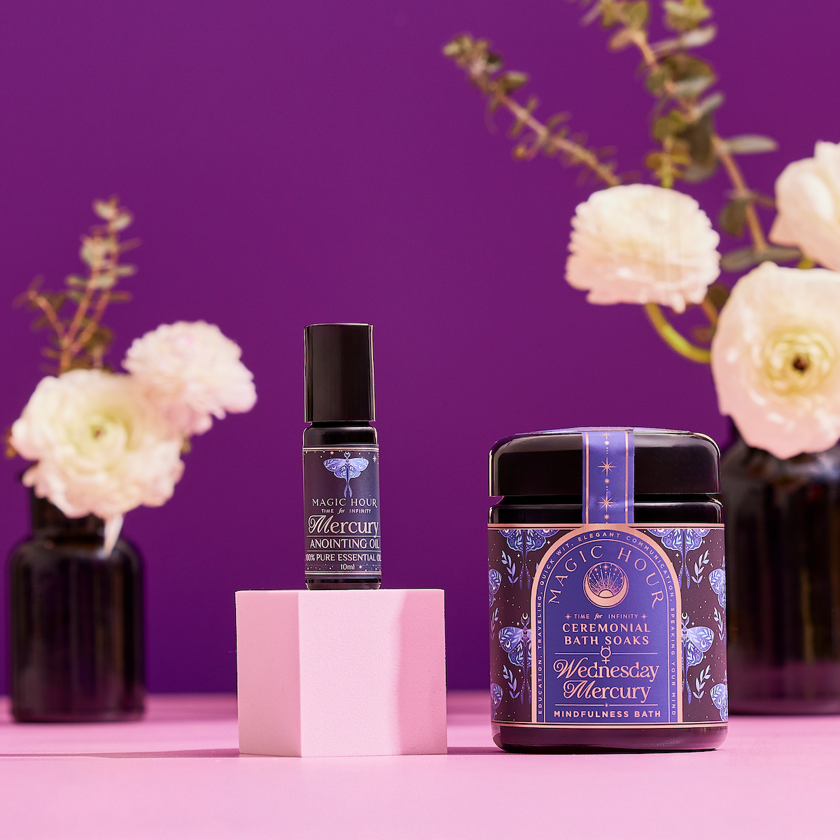 The Celestial Self-Care Ritual Set, featuring anointing oil and "Wednesday Mercury" bath soaks, is displayed on a pink surface with white flowers in black vases against a purple background, embodying planetary-inspired self-care.