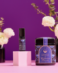 The Celestial Self-Care Ritual Set, featuring anointing oil and "Wednesday Mercury" bath soaks, is displayed on a pink surface with white flowers in black vases against a purple background, embodying planetary-inspired self-care.
