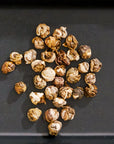 A cluster of dried jasmine flowers is displayed on a dark flat surface. The naturally curled round shapes, reminiscent of White Tea Pearls from Magic Hour, show variations of brown and beige hues, hinting at their exquisite taste.
