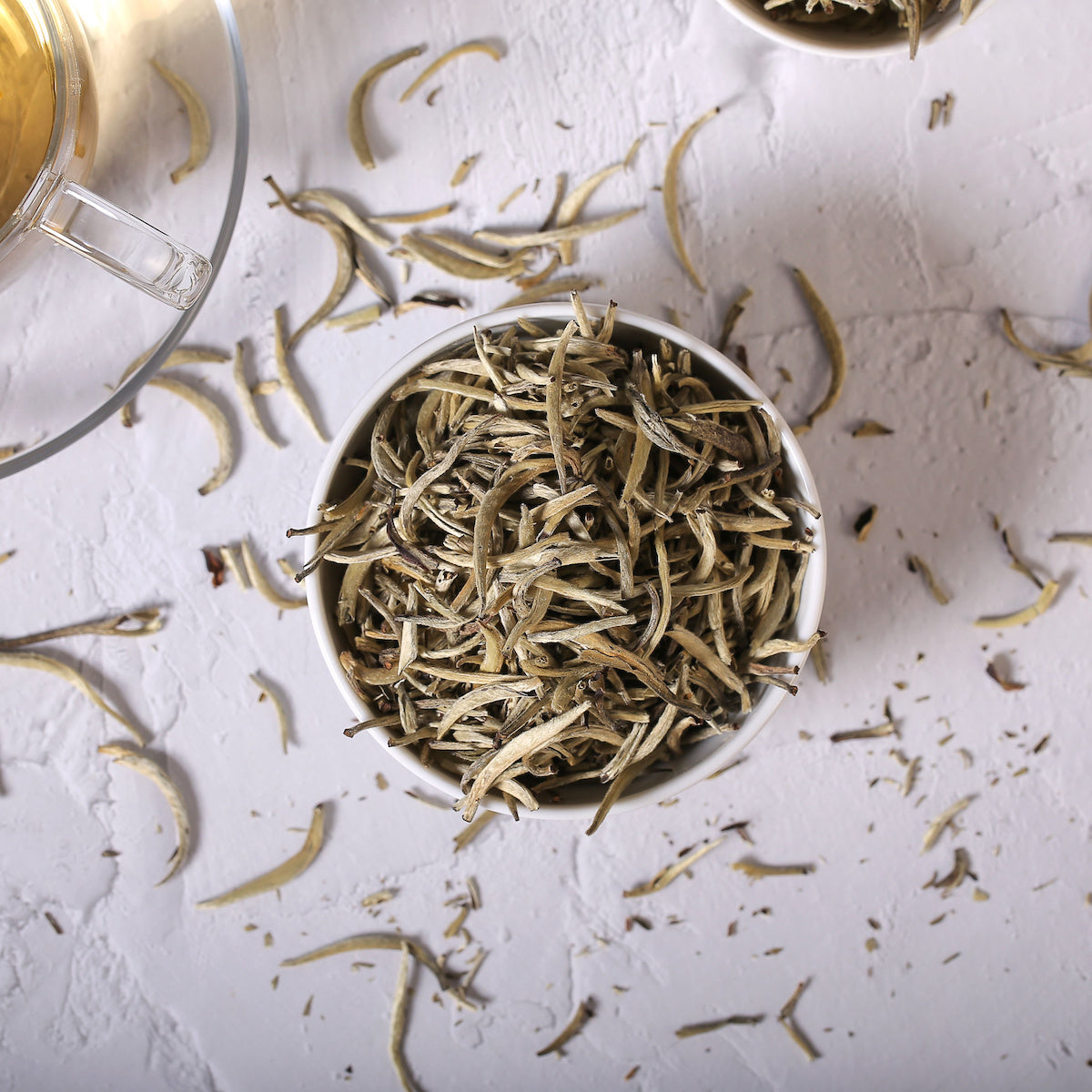 A bowl filled with dried white tea leaves is placed on a light-colored surface. Loose tea leaves are scattered around the bowl. Part of a glass teapot with brewed tea is visible on the left side of the image.