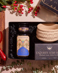 A flat lay of the "Winter Wellness Wishes Tea & Accessory Gift Set" features a luxury gift box containing a jar of seasonal teas, a ceramic tea bowl, and a pot on a wooden tray. It's beautifully complemented by red and yellow bread and decorative greenery with red berries.