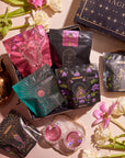 An assortment of elegantly designed tea packages promoting balance is displayed on a surface, surrounded by fresh flowers, a gold teapot, and a black box labeled "Magic Hour." Two pink tea cups and a small bowl with a white rim are also included in the Women's Wellness Sampler Set arrangement.
