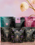 Six tarot card-inspired tea packages with celestial and floral designs are displayed on a pink surface amid white flowers—perfect for gifting or trying the Women's Wellness Tea Sampler Set, an organic blend crafted for wellness.