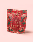 The "Year of the Horse: Adaptogen Chai Black Tea" comes in a red floral package with gold and pink text, decorative flowers, and vines—celebrating Lunar New Year tea against a pale pink background.