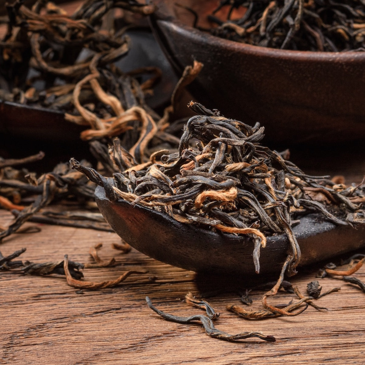 Close-up of Golden Monkey Yunnan Black Tea leaves with golden tips on a wooden spoon, scattered over a rustic wooden surface. The texture and details of this premium Chinese black tea are clearly visible.