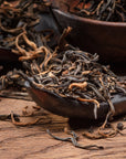 Close-up of Golden Monkey Yunnan Black Tea leaves with golden tips on a wooden spoon, scattered over a rustic wooden surface. The texture and details of this premium Chinese black tea are clearly visible.