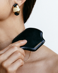 A woman with dark hair uses a black gua sha tool from the Beauty Inside & Out Box, inspired by Traditional Chinese Medicine, holding it to her neck while wearing gold and black earrings and a thin gold necklace.