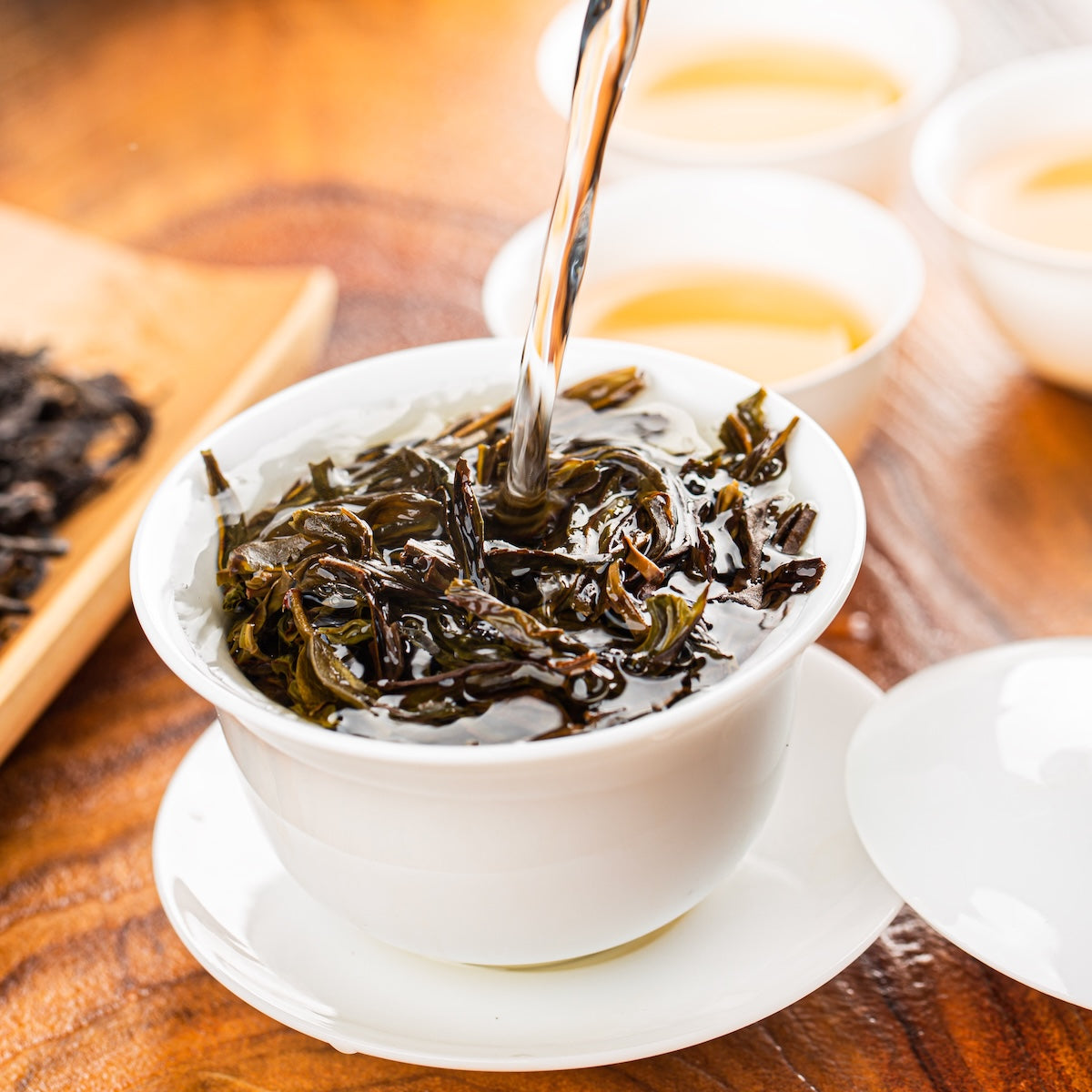Hot water is being poured over Zhangping Shuixian Oolong loose leaf tea in a white cup with a saucer, with more tea cups and a plate of loose Zhangping Shuixian Oolong leaves in the background.