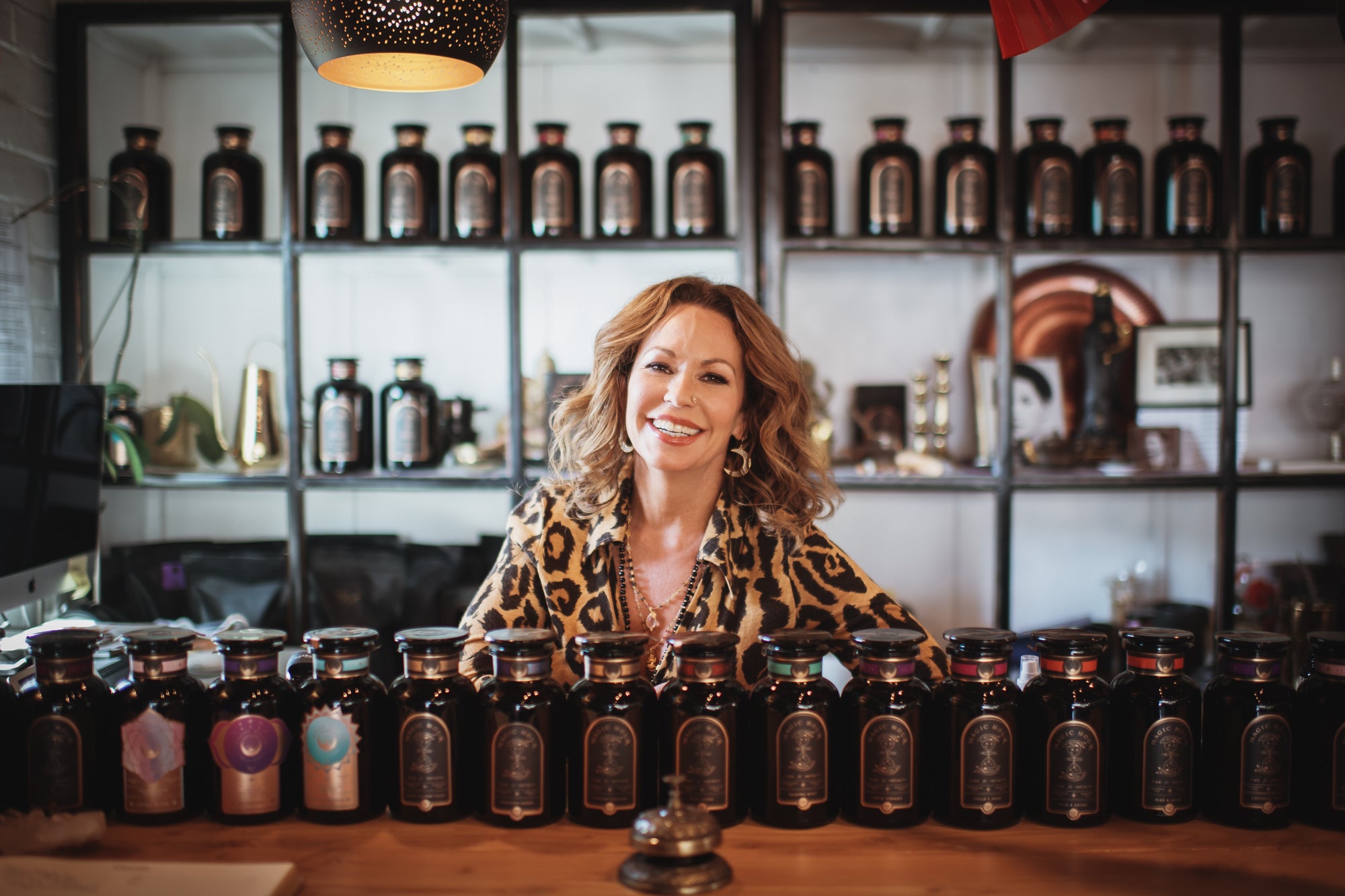 A woman with curly hair smiles while standing behind a counter with various dark-colored bottles of apothecary products displayed on it. Behind her, shelves filled with more bottles and various decor fill the background.