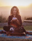 A woman sits cross-legged on a blue and brown patterned mat outdoors at sunset. She holds a teacup with both hands, and in front of her are a glass teapot and a jar on a wooden tray. She has long hair and wears a dark robe. The background is a serene, mountainous landscape.