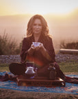 A woman sits cross-legged on a blue and brown patterned mat outdoors at sunset. She holds a teacup with both hands, and in front of her are a glass teapot and a jar on a wooden tray. She has long hair and wears a dark robe. The background is a serene, mountainous landscape.