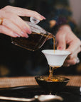 At the Ojai Tea Salon, a person pours tea from a glass teapot into a small white and black teacup, surrounded by Tea Essentials and the In Person & Virtual Tea School Course. Steam rises as a spoon rests nearby.