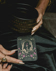 A person with manicured nails holds a black cup adorned with gold stars and displays “The Lovers” card from the Magic Hour 12 Archetype Set on their lap, dressed in a black satin outfit and wearing statement rings.