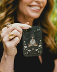 A woman with wavy hair holds up a tarot card titled "The Oracle." The card features an illustration of a figure with flowing hair, sitting in a meditative pose, surrounded by lilies, with celestial symbols above. The woman smiles, with greenery in the blurred background.
Magic Hour