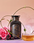 An Aries Astrology Tea Gift Set featuring an apothecary jar labeled “Aries” next to a glass cup of tea, a pink rose, and blackberries.