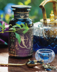 A glass teapot with blue tea, a small glass cup, and a brass teaspoon of loose tea complement the "Monthly Magic Tea Journey Subscription," creating a goddess ritual celebrating the Divine Feminine amid lush greenery.