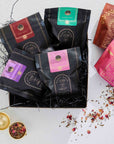 The Herbal Tea Sampler Tea Box offers caffeine-free teas for sleep, digestion & immunity, beautifully arranged with a gold tea strainer and rose petals on black shred, plus two extra tea bags on a white surface.