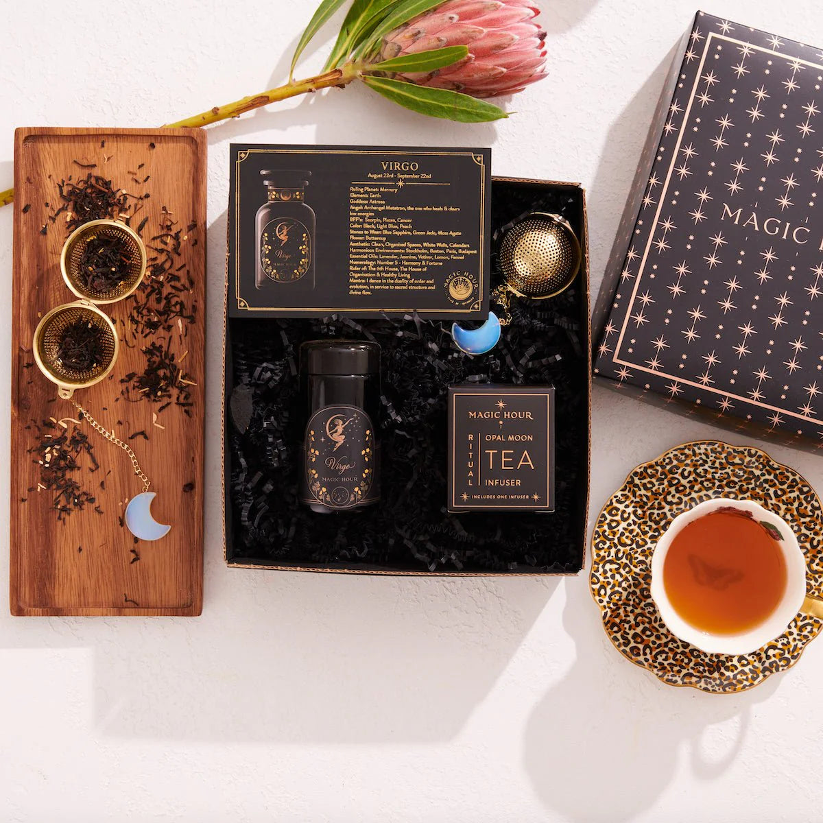 Flat lay of the Virgo Astrology Tea Gift Set, showcasing a decorative box labeled "Magic." The set includes a jar of loose tea made with organic ingredients, a card labeled "Virgo," a tea infuser, and a wooden tray adorned with moon-shaped charms. Enhance your ritual by enjoying this wellness blend and sip of tea.