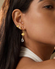 A woman with long dark hair, wearing a white sleeveless top, gazes thoughtfully while showcasing the Freedom to Soar Butterfly Stud Earrings—18k gold plate studs featuring a teardrop gemstone and a cluster of small butterflies.