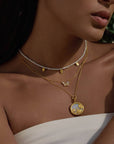 A woman wears the Renewed Hope Pearl Butterfly Necklace—an 18k gold plated multi-layer piece featuring pearls, small gold charms, a butterfly, and a round locket—styled perfectly with a white off-shoulder top.