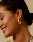 A woman with dark hair in a low bun smiles with her eyes closed, wearing multiple gold earrings—including a large textured hoop, Inner Light Blue Topaz Pear Stud Earrings, and smaller studs and cuffs. She wears a light-colored top.