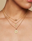 A woman wears a 16" Emerald Dot Cross Pendant Necklace with a 3" extension, styled with other gold layers and a light-colored top, highlighting her elegant faith-inspired jewelry look.