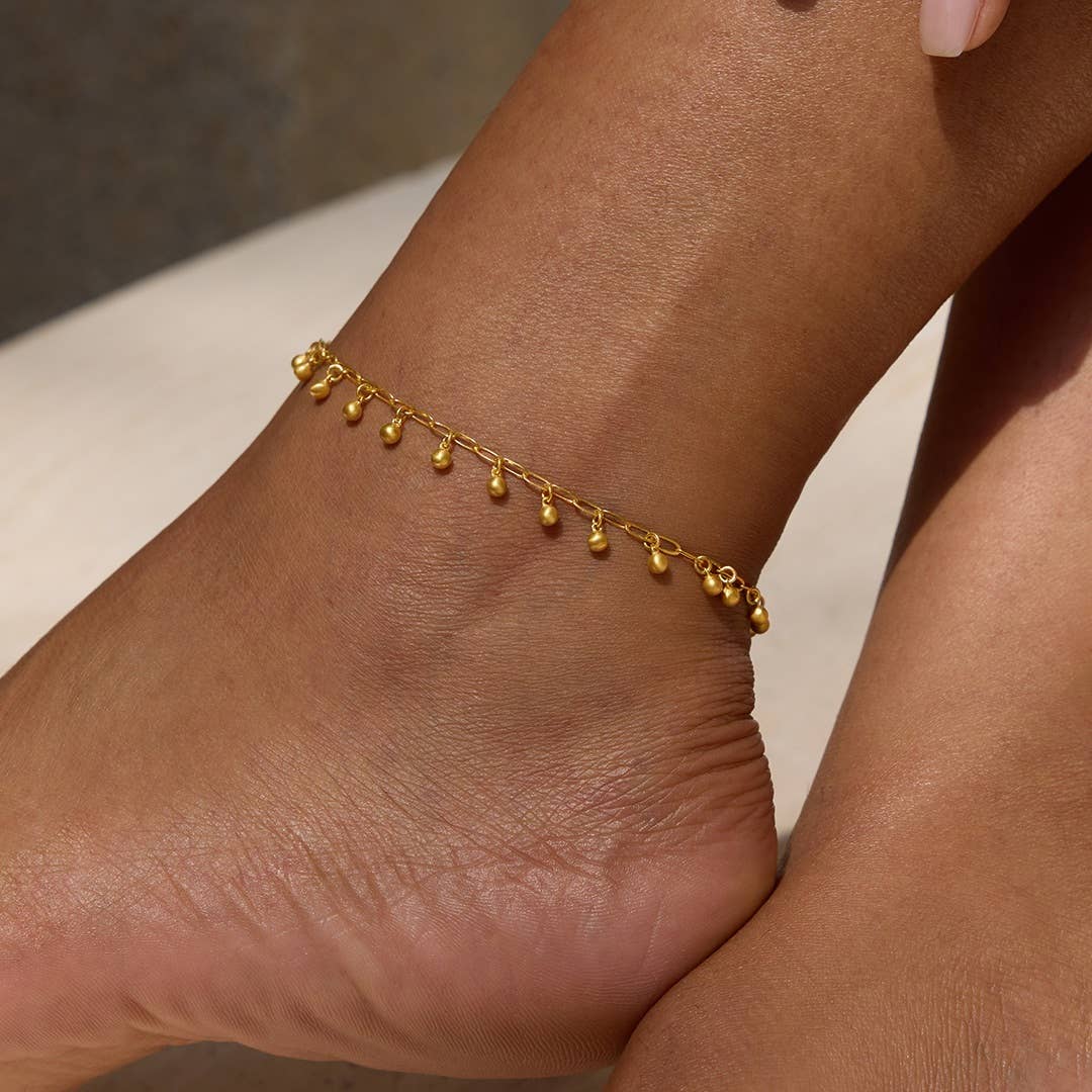 The Joyful Anklet, crafted on an 18k gold plated oval cable chain with delicate dangling charms, graces the ankle in this close-up against a neutral background, highlighting its elegant design.