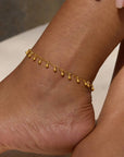 The Joyful Anklet, crafted on an 18k gold plated oval cable chain with delicate dangling charms, graces the ankle in this close-up against a neutral background, highlighting its elegant design.