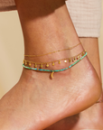 A close-up shows a person's ankle wearing three anklets: a delicate gold chain, a gold chain with small charms, and the 9" Turquoise moon anklet with beads, a small gold charm, and a 1" extender. The background is soft and neutral.