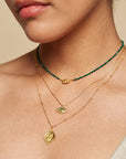 A close-up of a woman’s neck shows her layering necklaces, including the 16" Emerald Hamsa Pendant Necklace with a 3" extension chain. She pairs it with a green beaded choker, an evil eye gold chain, and wears a light top.